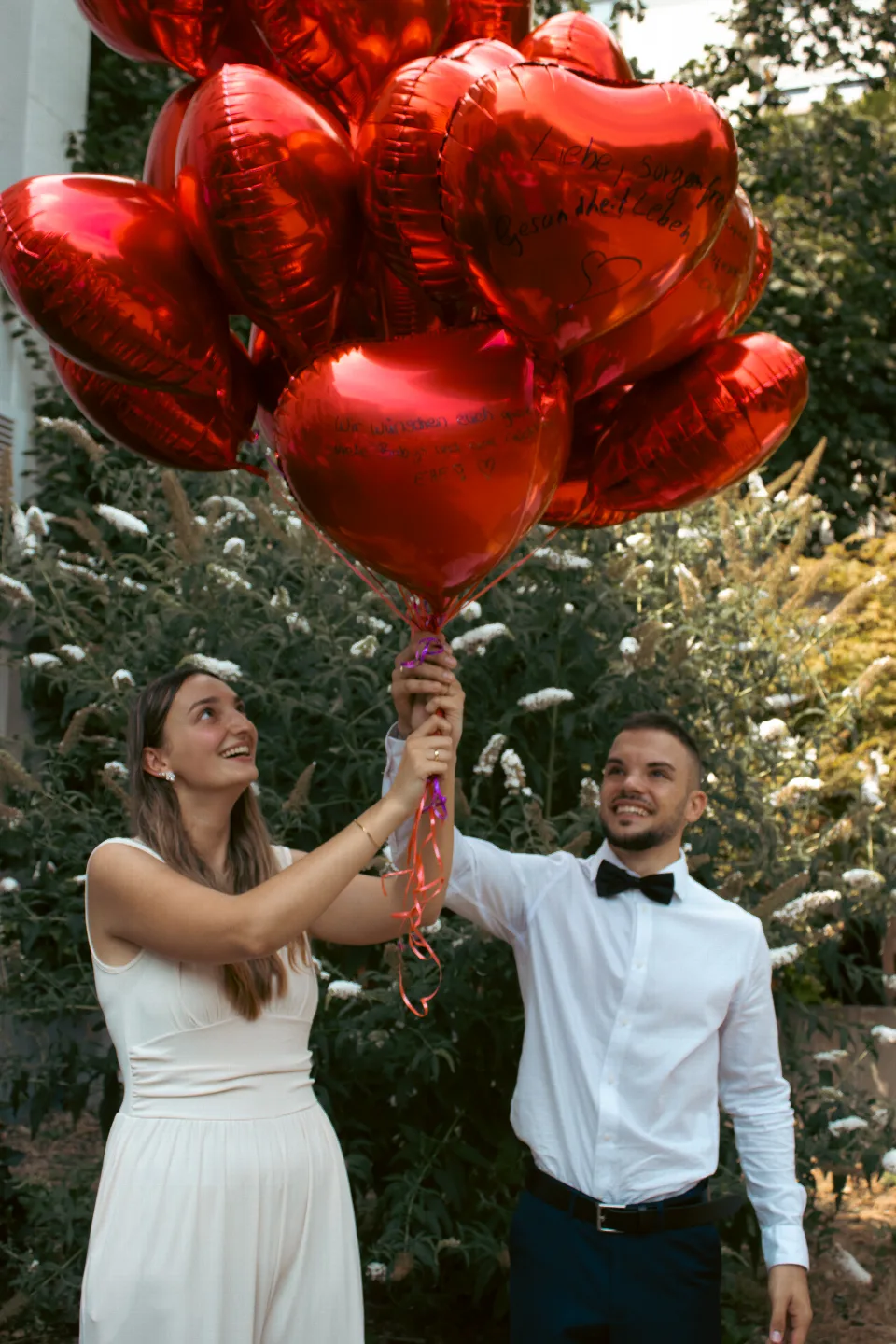 Brautpaar in festlicher Kleidung hält auf dem Gelände des Standesamtes gemeinsam rote, herzförmige Luftballons mit Wünschen der Hochzeitsgäste.