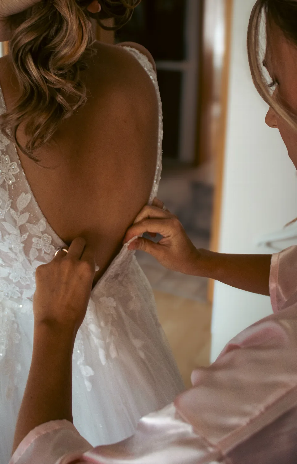Nahaufnahme einer Braut im Spitzenbrautkleid mit tiefem Rückenausschnitt, während eine weitere Person das Kleid vor der Hochzeit zuknöpft.