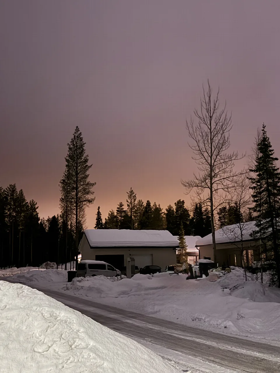 Verschneite Häuser und Fahrzeuge vor schneebedeckten Nadelbäumen in der Dämmerung in Ylläsjärvi, Finnland.
