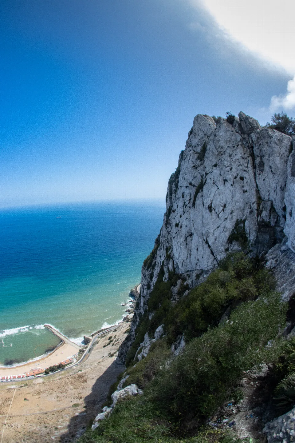 Felsformation mit steiler Klippe und grüner Vegetation am Rand, darüber das Meer mit einem Strand und Hafen im Hintergrund bei Gibraltar.